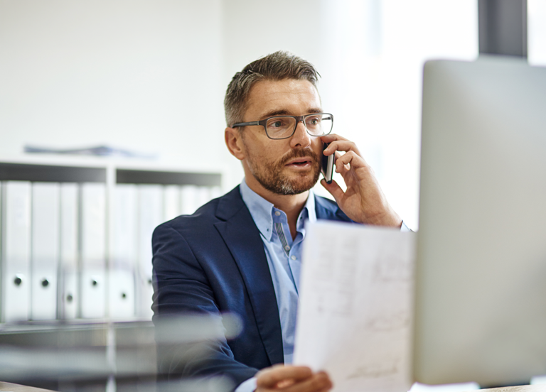 Businessman using phone and computer while reading a document at his desk